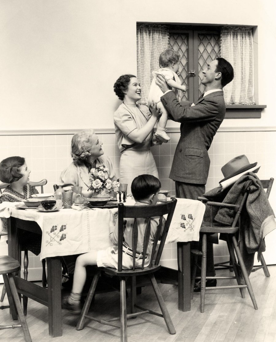 Photographie sépia d'une famille autour d'une table, un père soulevant un nourrisson.